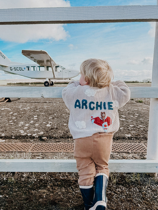 A blonde toddler wearing a jumper with the name Archie stands in front of a light aircraft