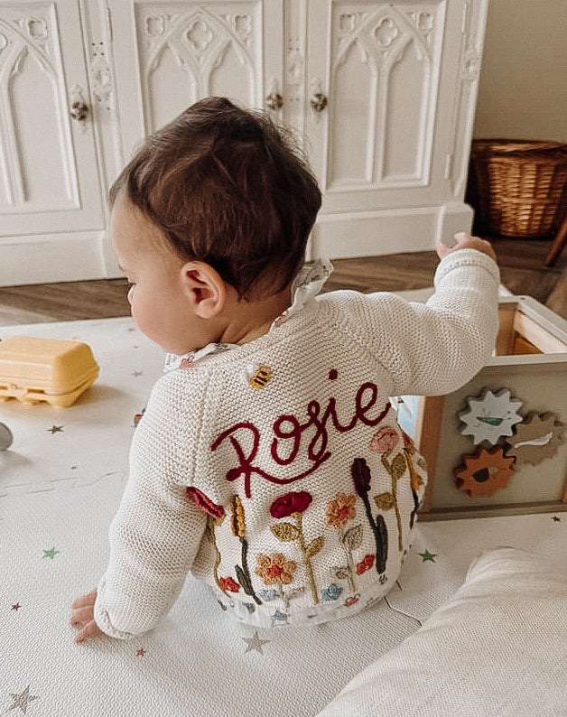 A toddler wearing a white cardigan with the name Rosie on the back